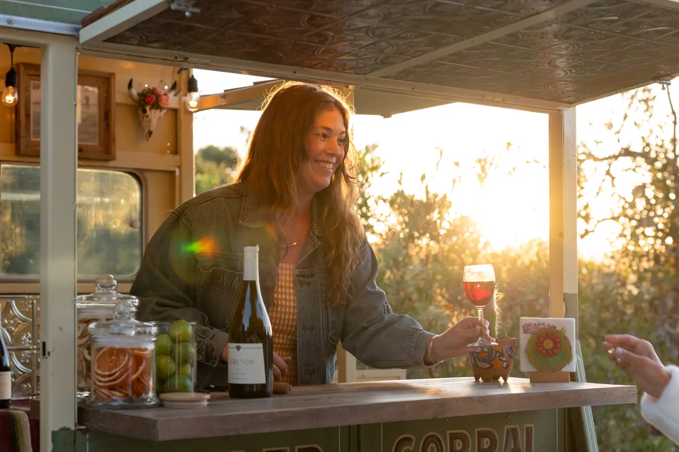 A bartender hands a glass of red wine across the bar as the sun sets behind the trailer